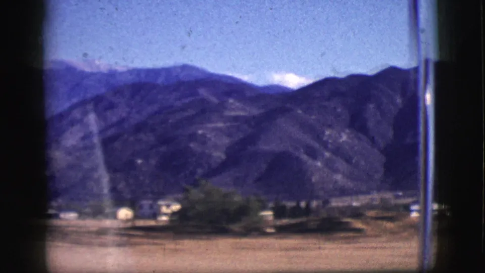 The 8mm frame shows a low valley, scattered roadside buildings, trees, and mountains beyond a streaked vehicle window.