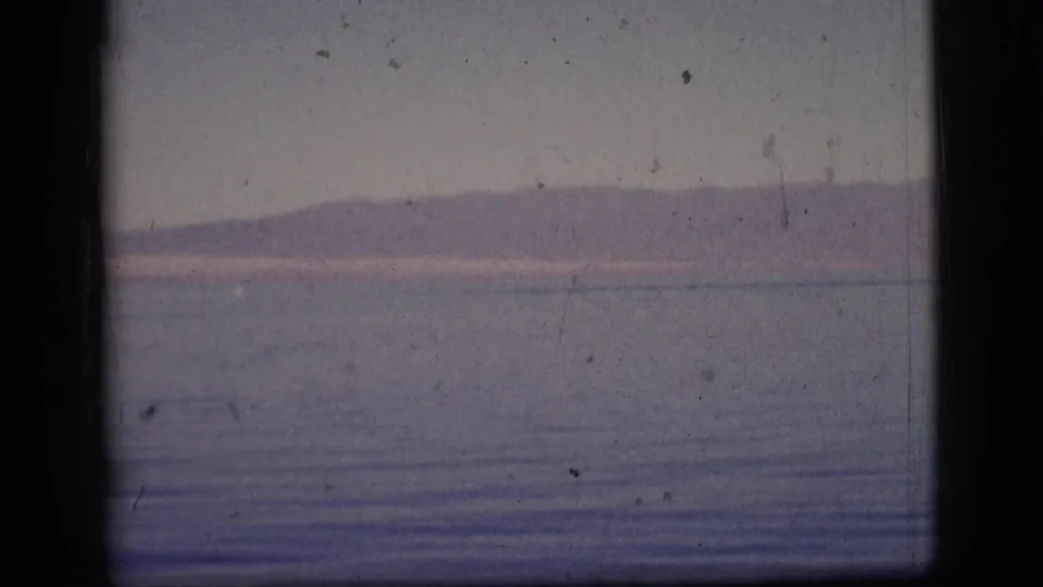 1961 8mm frame showing a low mountain ridge beyond still water, with faded blue-purple color and small film scratches.
