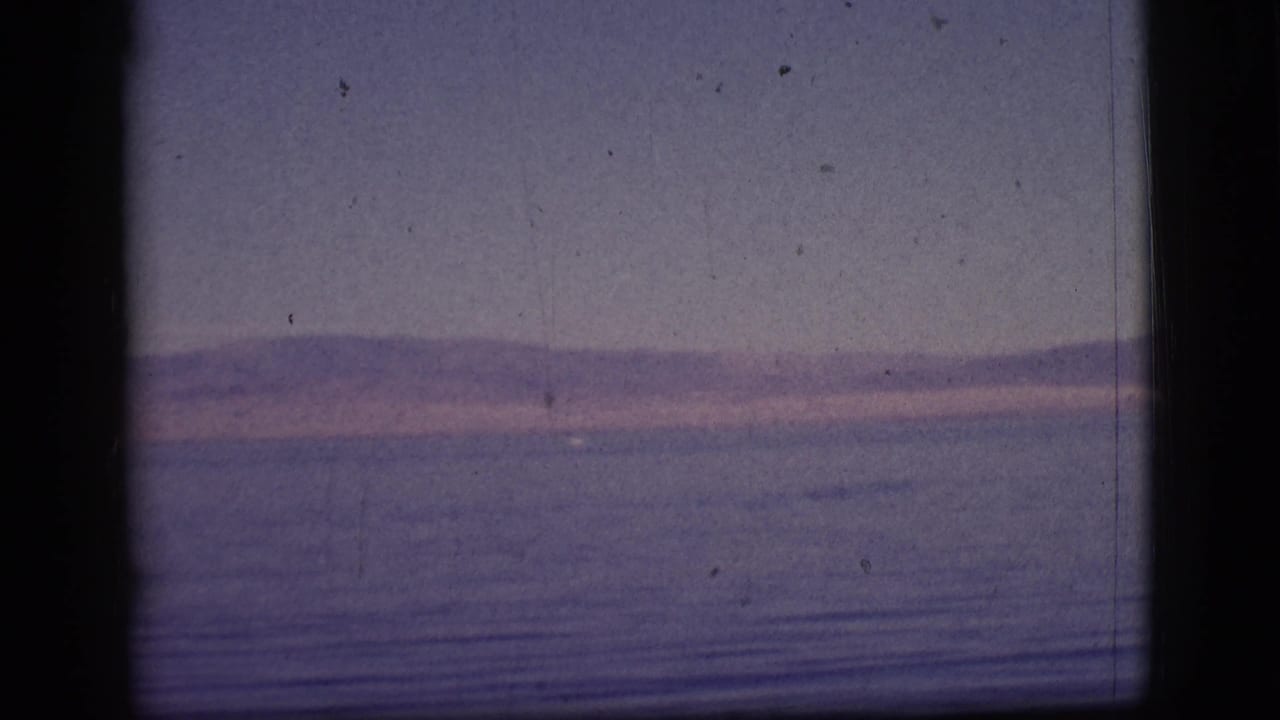 Broad blue water stretches toward low hills beneath a softly colored sky and visible film scratches.