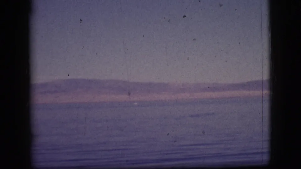 A vintage 8mm frame shows a broad water surface, low mountains, and pink-purple sky within the scanned film gate.