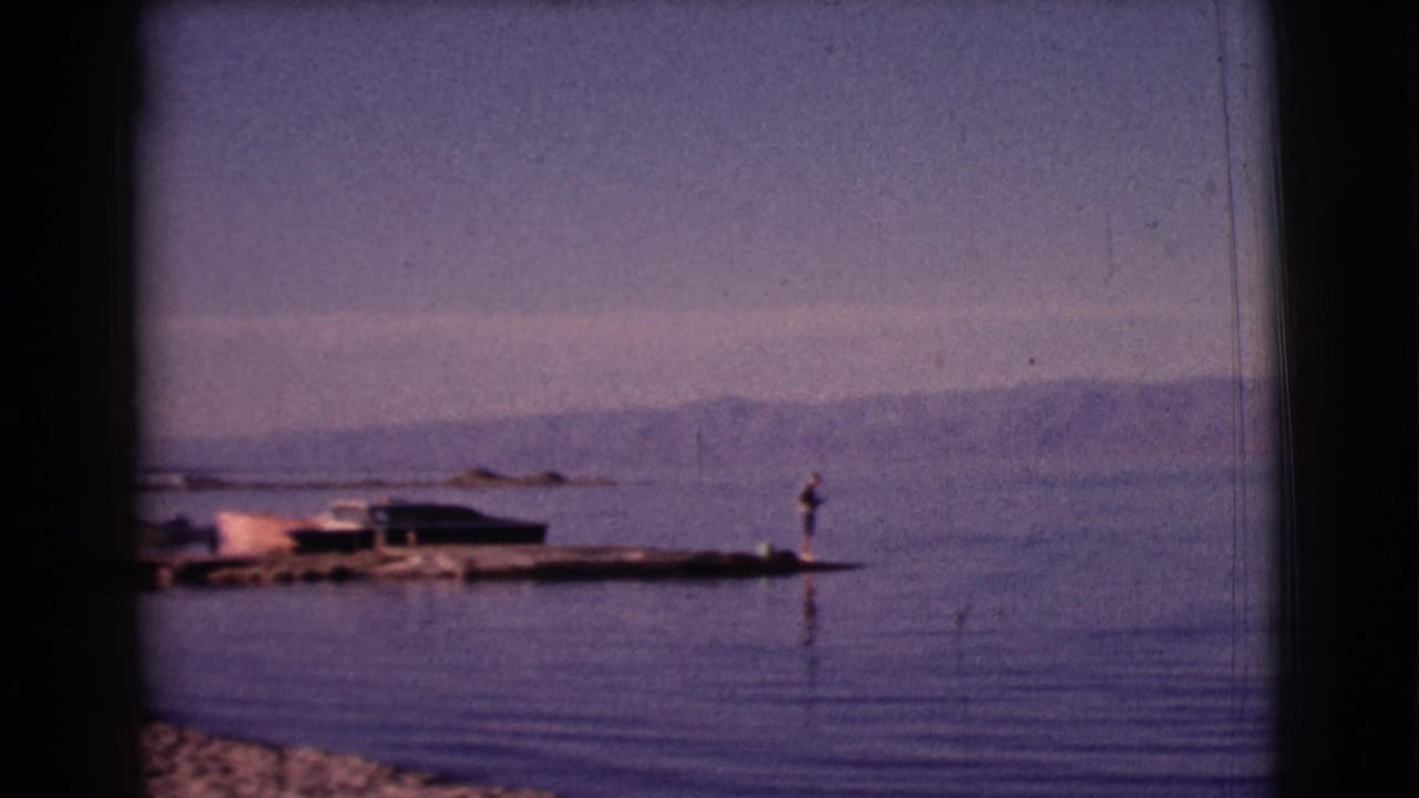 A calm lake with a small dock, fishing figure, boats, and distant mountains.