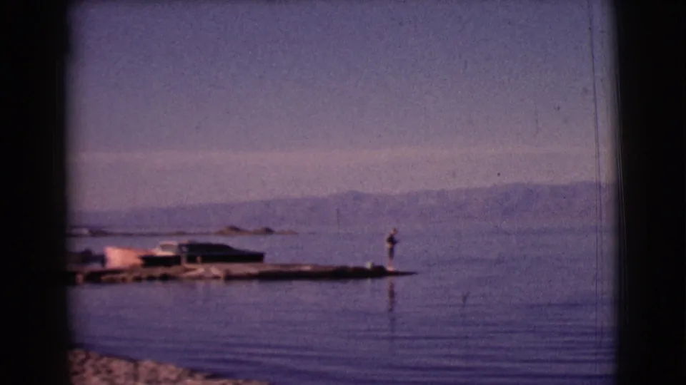 1961 Clarkdale, Arizona home-movie frame showing a lake, small dock, lone fishing figure, boats, and hazy mountains.