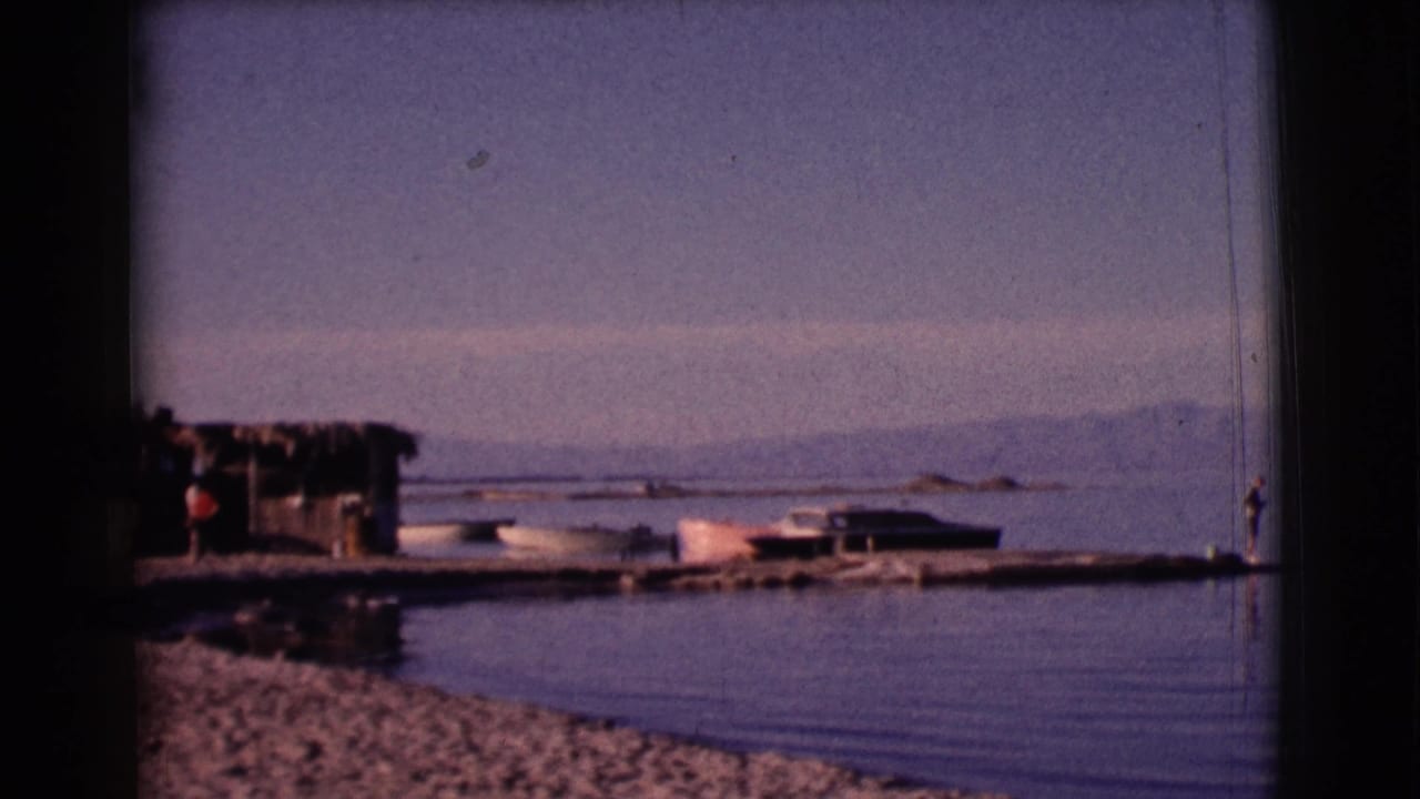 A lakeshore with a rustic shelter, small boats, gravel bank, and mountains beyond.