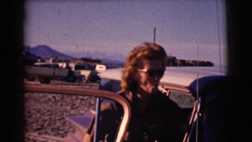 1961 Clarkdale, Arizona frame showing a woman in sunglasses smoking beside an open car door near boats and a lakeshore.