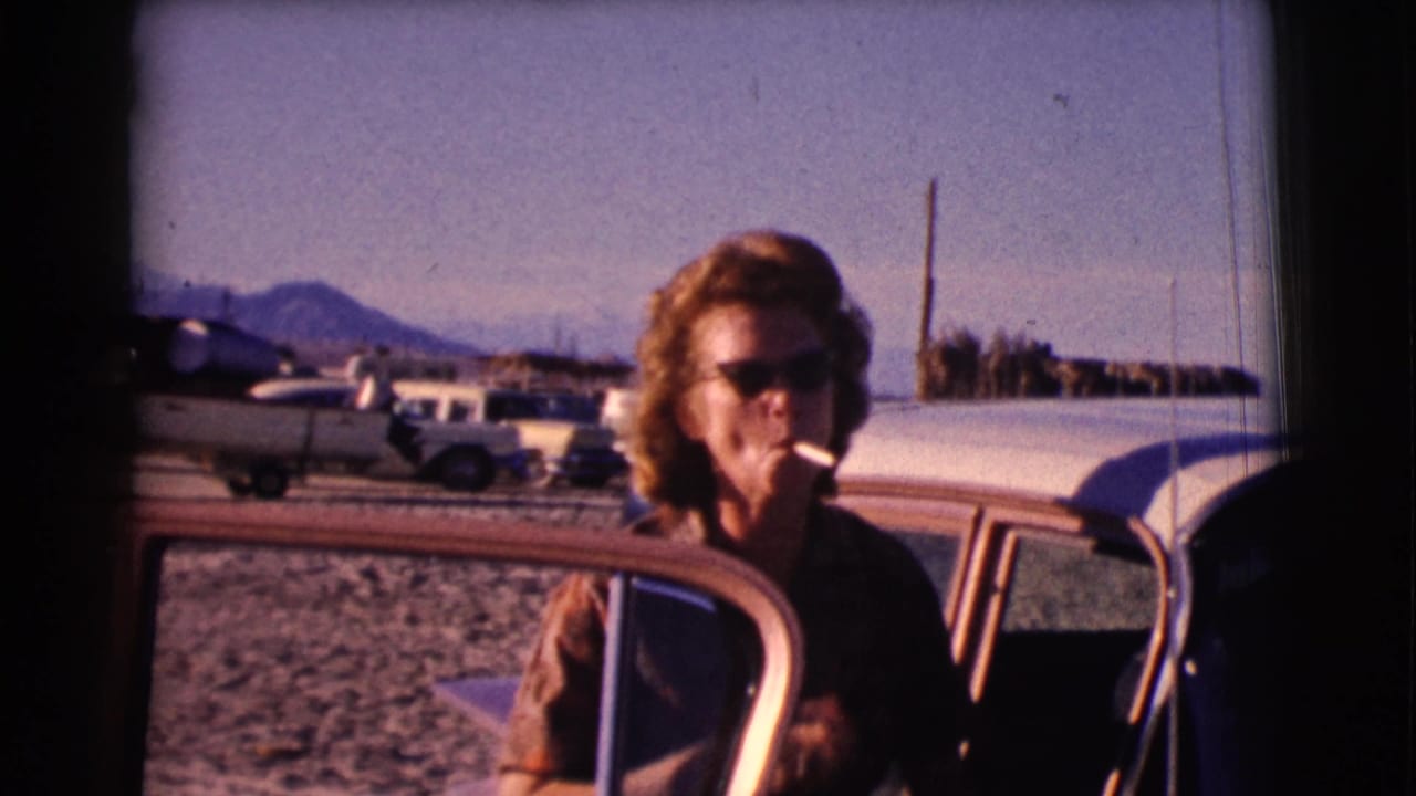 Woman in sunglasses smoking beside an open car door in a dusty lot.