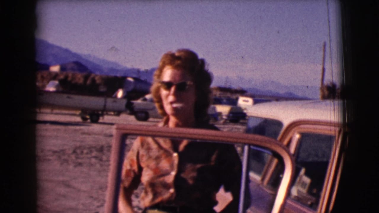 Woman standing by an open car door with parked vehicles and mountains behind her.