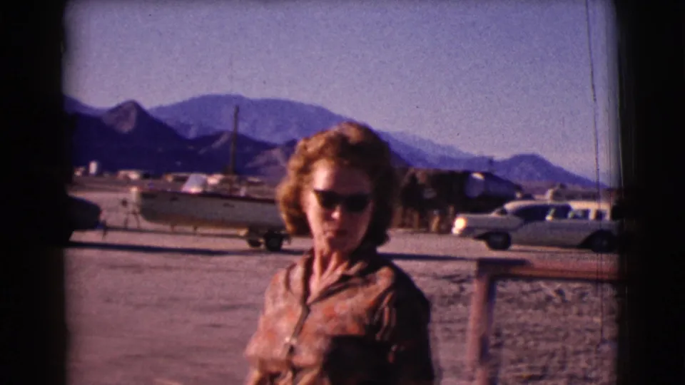 In the 1961 Clarkdale home movie, the woman faces the camera with cars, a boat on a trailer, and mountains across the dusty lot.