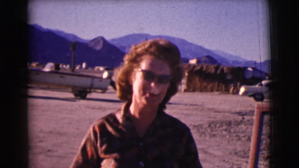 Adult woman in sunglasses leans slightly near a car door in Clarkdale, Arizona, with a boat trailer, cars, and mountains behind her.