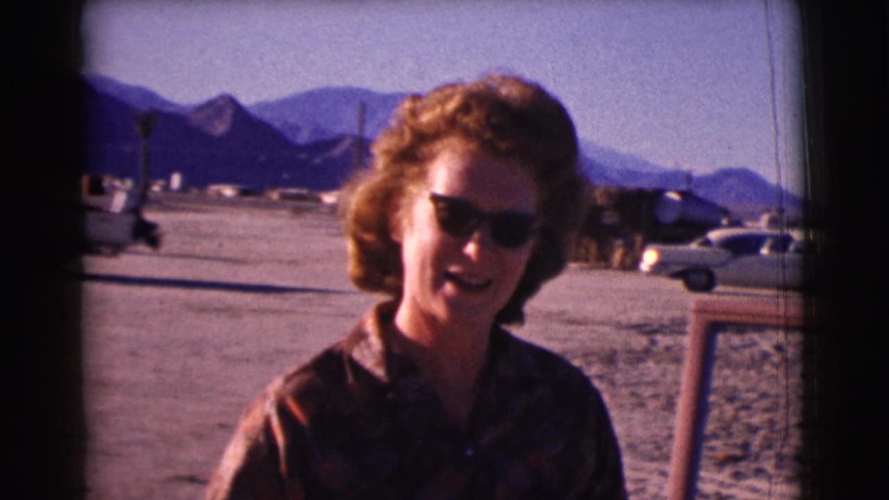 Woman in sunglasses faces the camera near parked cars with mountains behind her.