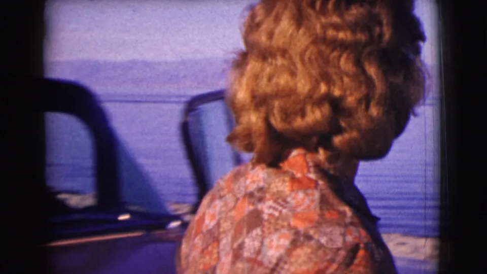 The woman is seen from behind near a car, turning toward the water and distant hills in the Clarkdale, Arizona home movie.
