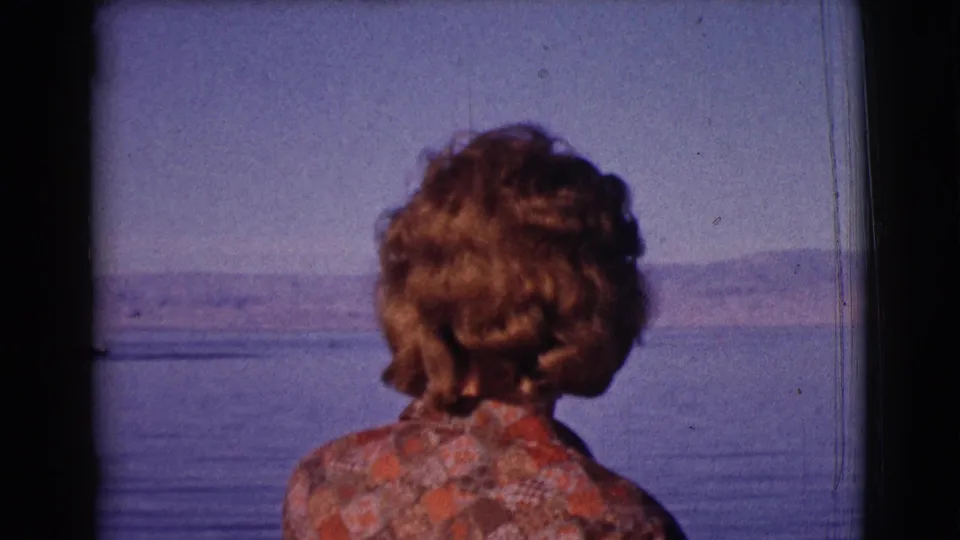 From behind, the woman faces a calm body of water and distant hills in a brief 1961 Arizona travel scene.