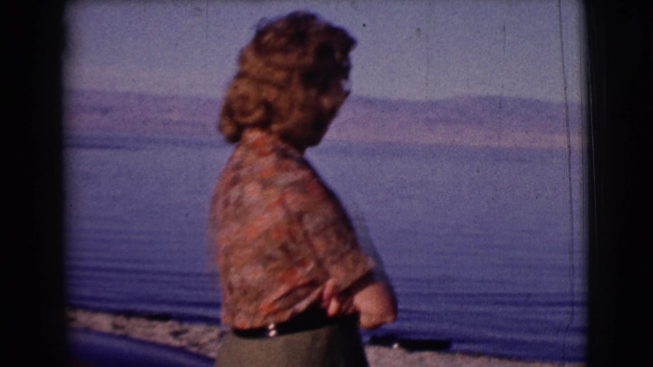 Woman in profile stands near calm water and distant hills.