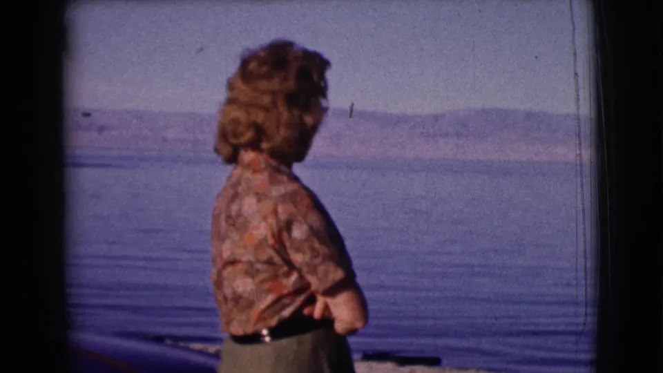 1961 8mm frame of a woman in side view beside a broad body of water, with hills visible beyond the surface.