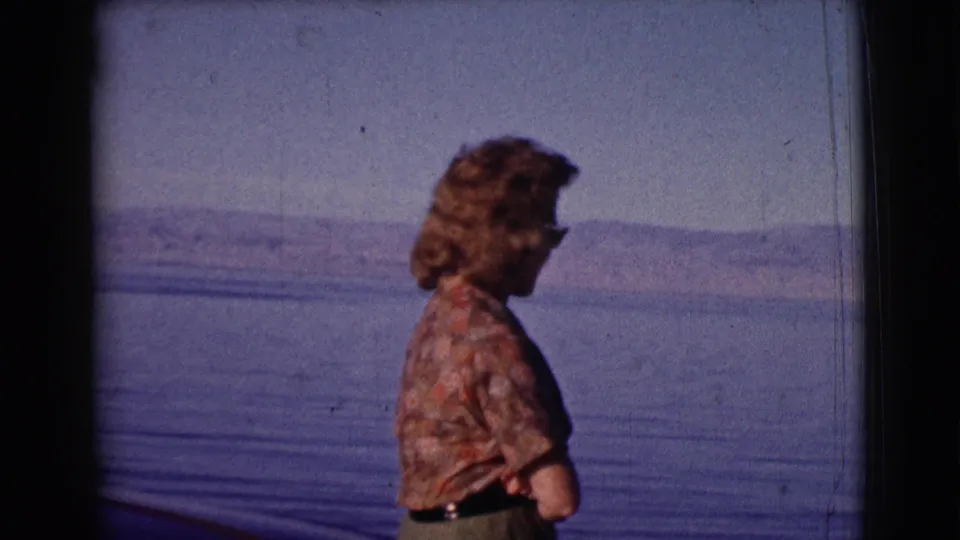 1961 8mm frame with the woman in profile, sunglasses visible, standing before blue water and low distant hills.