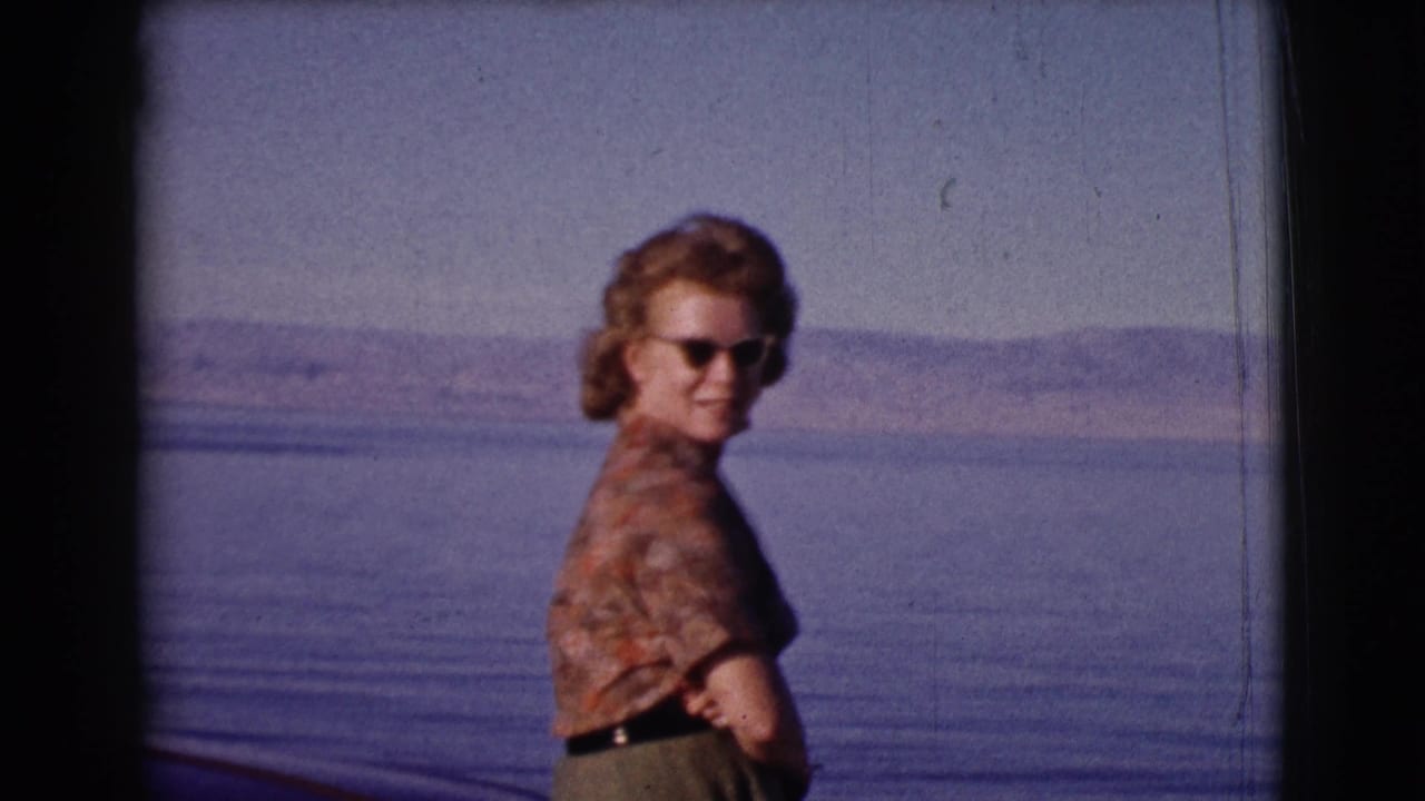 Woman in sunglasses stands beside water with hills in the distance.