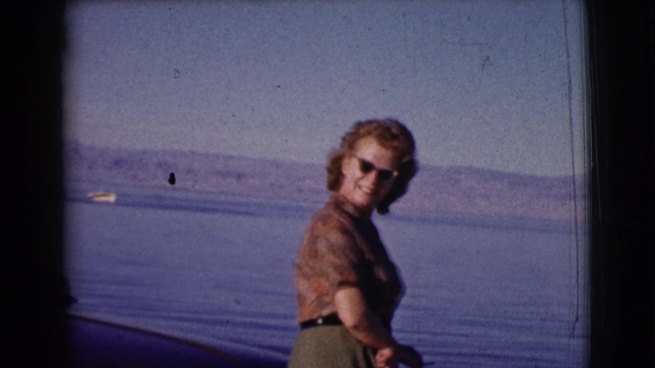 Woman smiles toward the camera beside water with a small boat in the distance.
