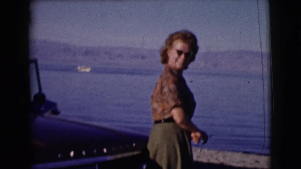 A woman in sunglasses stands beside the front of a vintage car near water, with a boat and hills in the background.