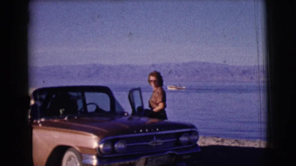 The woman stands beside a vintage car with an open door near the water, with a small boat and hazy hills beyond.