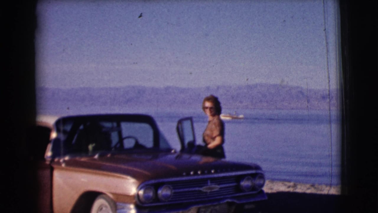 Woman standing beside a red convertible near a lake with mountains in the distance.