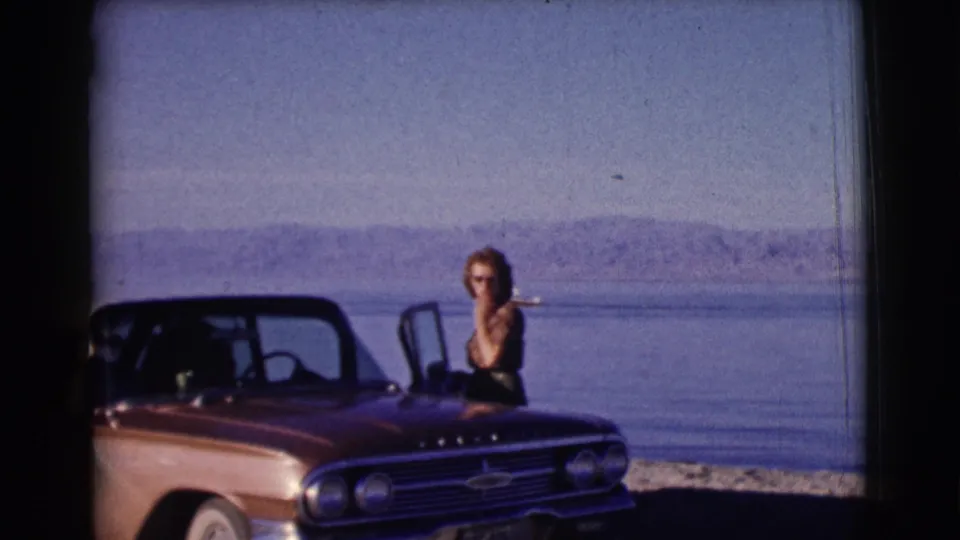 A woman poses at the open door of a red Chevrolet convertible near a calm Clarkdale lake, with a small boat and hills in the background.
