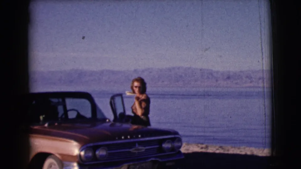 The woman leans near the front fender of a red Chevrolet convertible on a lakeshore in archival 1961 Clarkdale, Arizona home-movie footage.