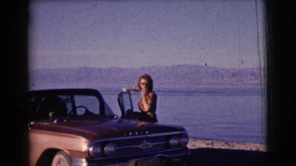 A woman in sunglasses stands beside the open door of a red Chevrolet convertible, framed by lake water and distant hills in Clarkdale, Arizona.