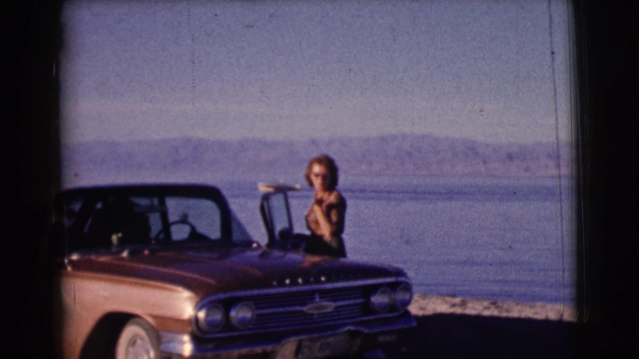 Woman in sunglasses stands beside a brown convertible near a lake and distant hills.