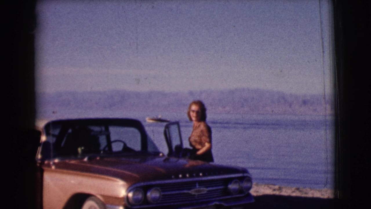 Woman stands by the open door of a brown convertible with water and hills in the background.