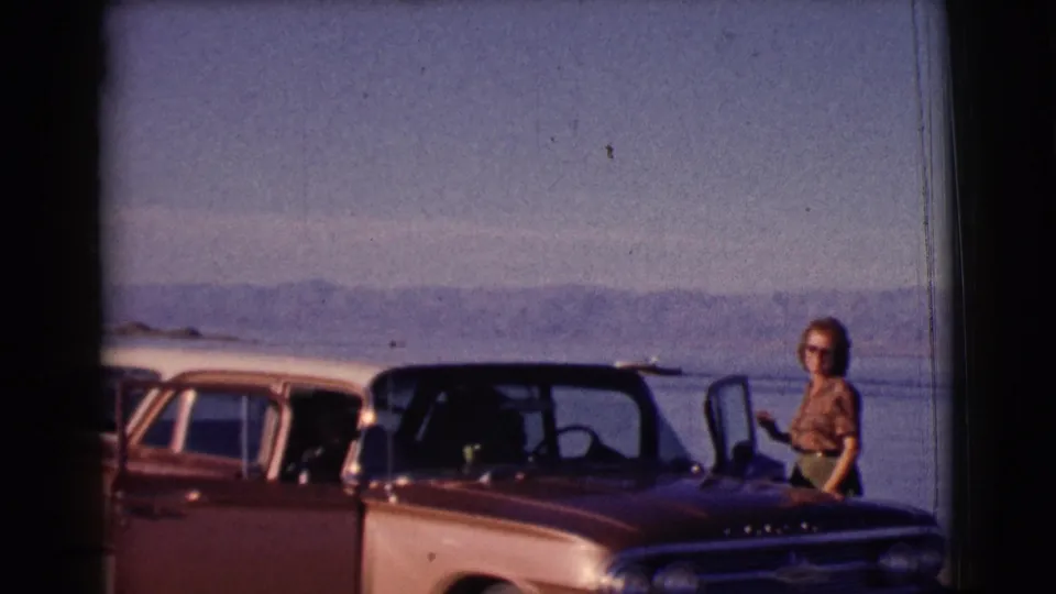 A second two-tone car enters the frame while the woman stands near the brown convertible at the lakeside beach.