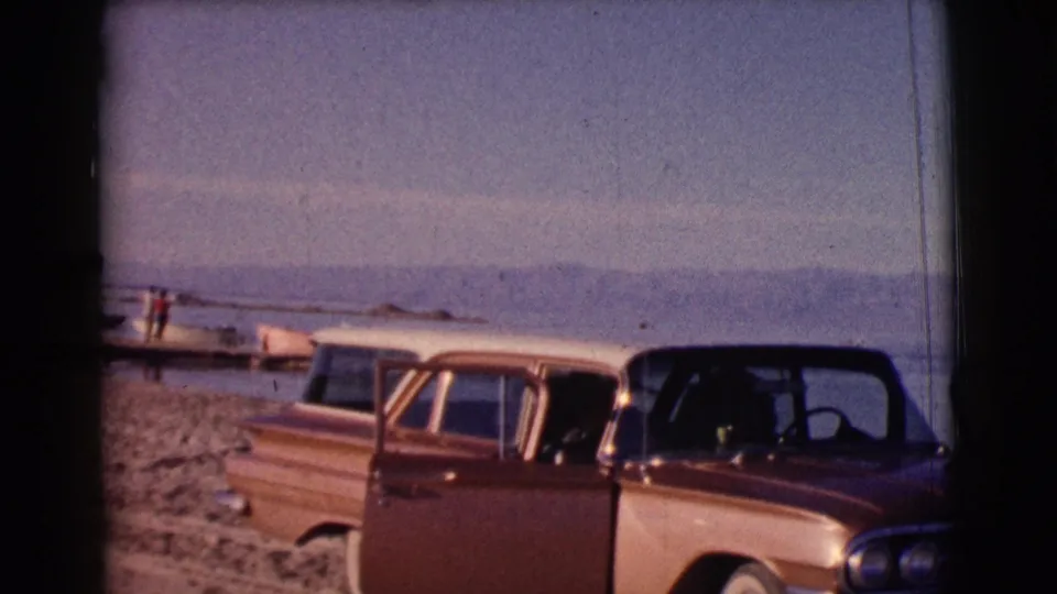 The final frame shows the parked cars from a wider angle, with beach, lake, distant hills, and a small boat visible behind them.