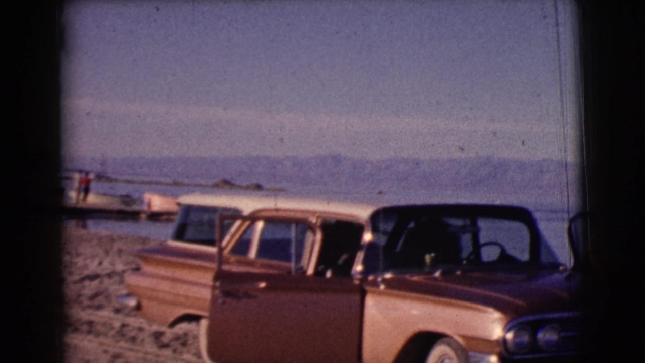 Station wagon parked on a sandy lakeshore with water and mountains behind it.