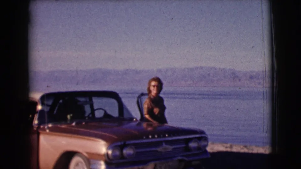 1961 Clarkdale, Arizona frame showing a woman in sunglasses beside the front of a station wagon with lake water behind her.