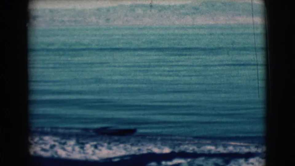 1961 Clarkdale, Arizona 8mm frame showing calm lake water, low waves, rocky shoreline, hazy hills, and a small distant boat.