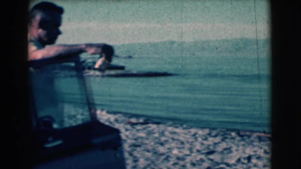 Adult man in a light shirt leans near a vehicle window frame at a waterside location in 1961 footage identified as Clarkdale, Arizona.