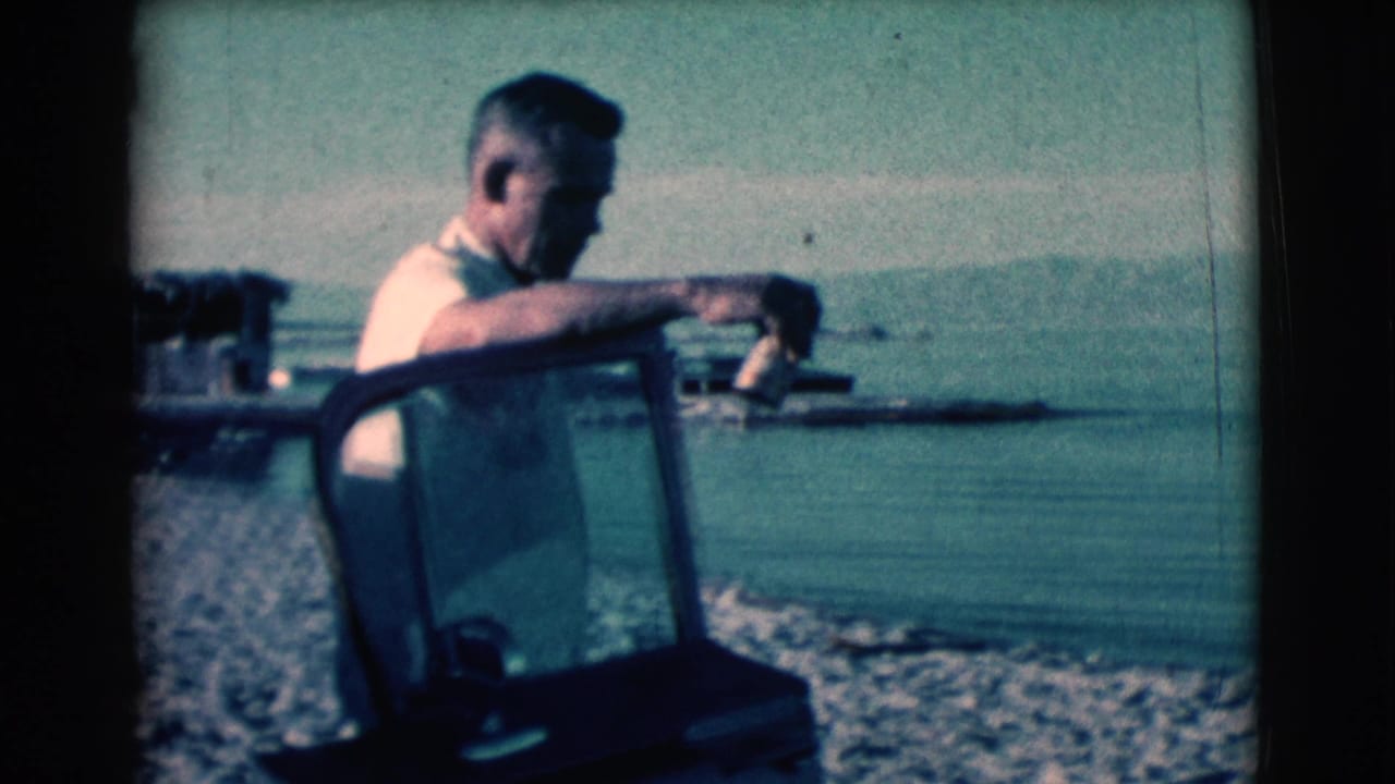 Man in a light shirt resting his arm on a vehicle frame beside a calm body of water.
