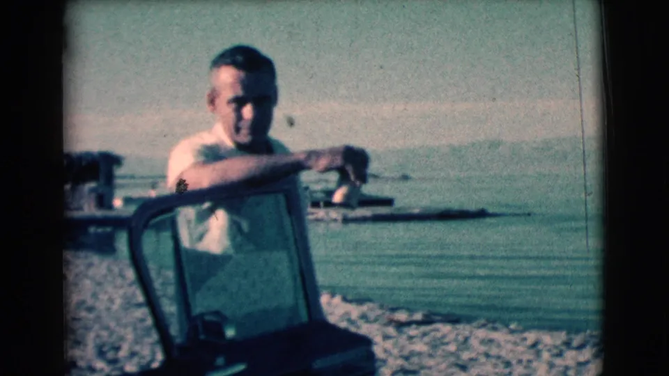 Adult man faces the camera while leaning on a vehicle window frame, with water and shoreline behind him in the 1961 clip.