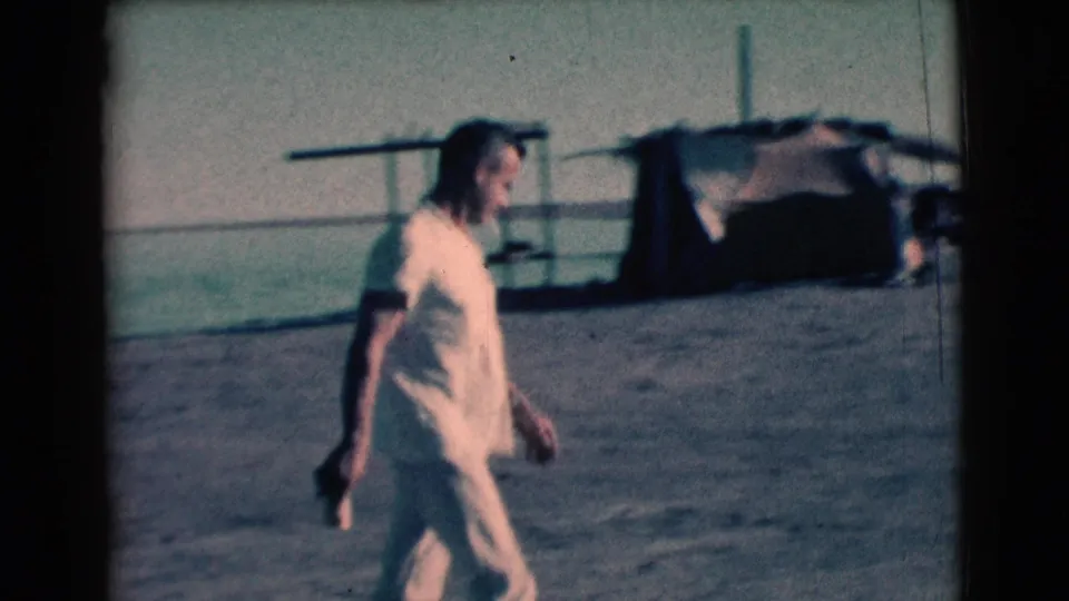Adult woman in light clothing walks across a beach or shoreline area near water and a small structure in the same 1961 home-movie scene.