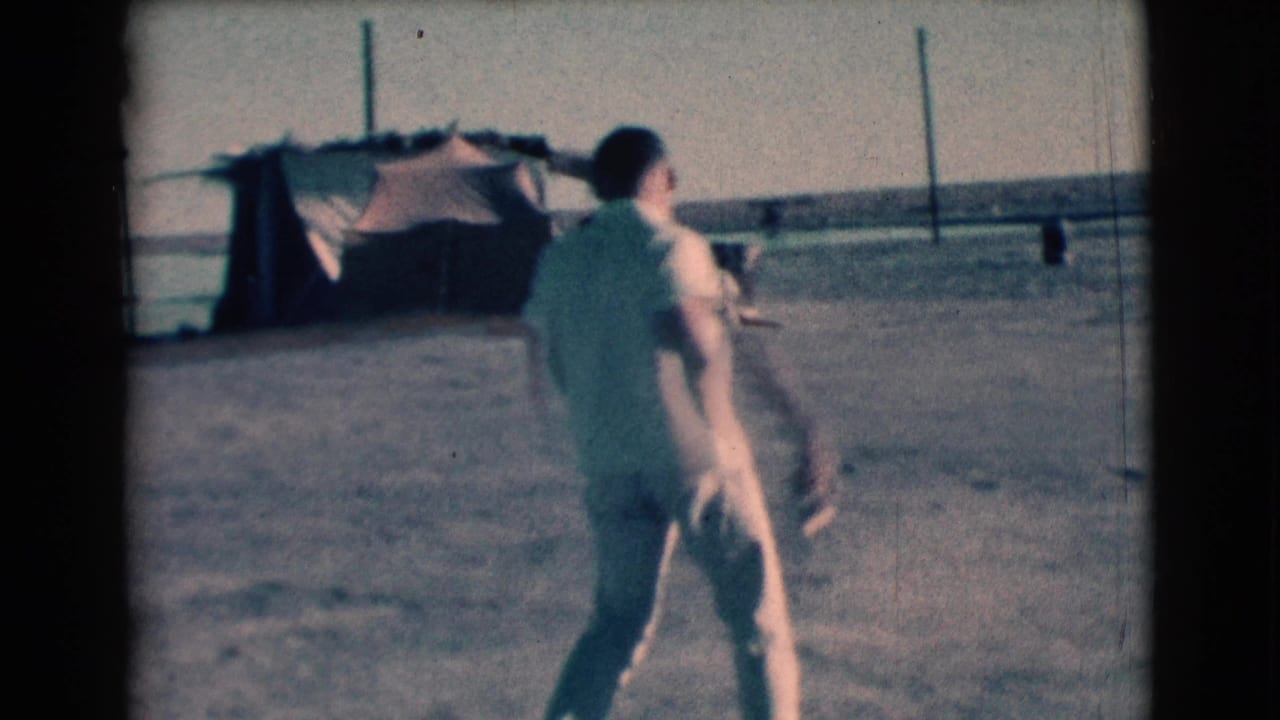 Man in light clothing walks across a dusty open area near low structures and fence posts.