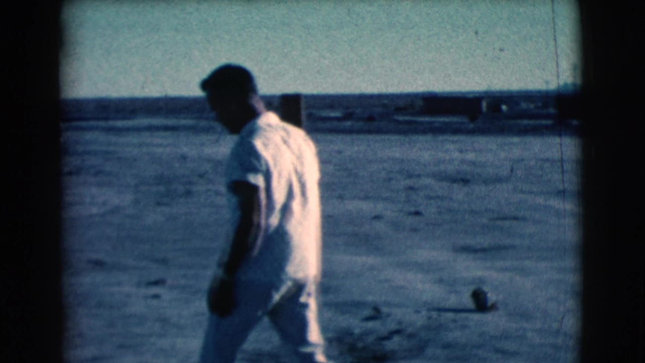 A man in a white shirt and light trousers walks across a dusty Clarkdale field, with fence posts and open sky in the distance.