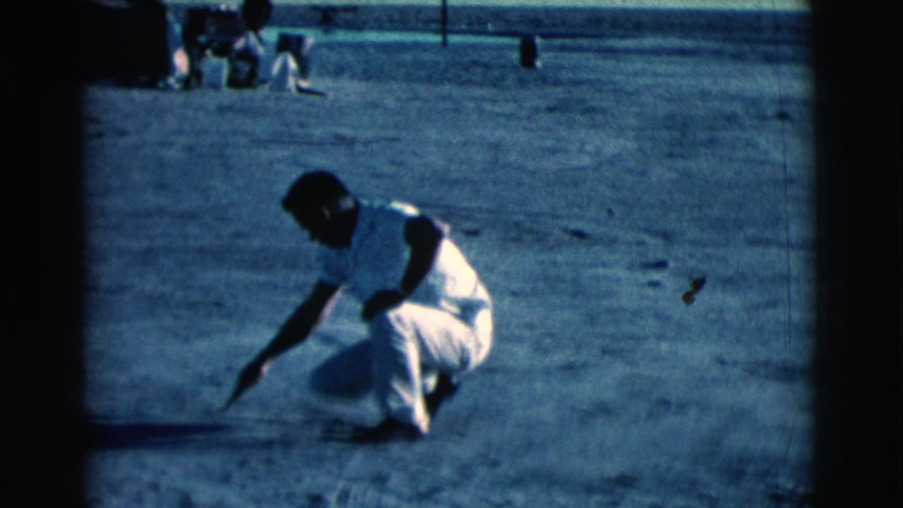 Man in white clothing squats on a dirt field and reaches down with a stick.