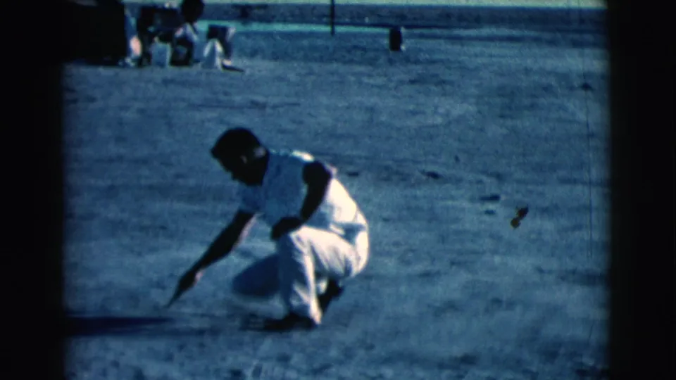 In Clarkdale, Arizona in 1961, a man in a white shirt and trousers squats in bright daylight and touches a stick to the dirt.