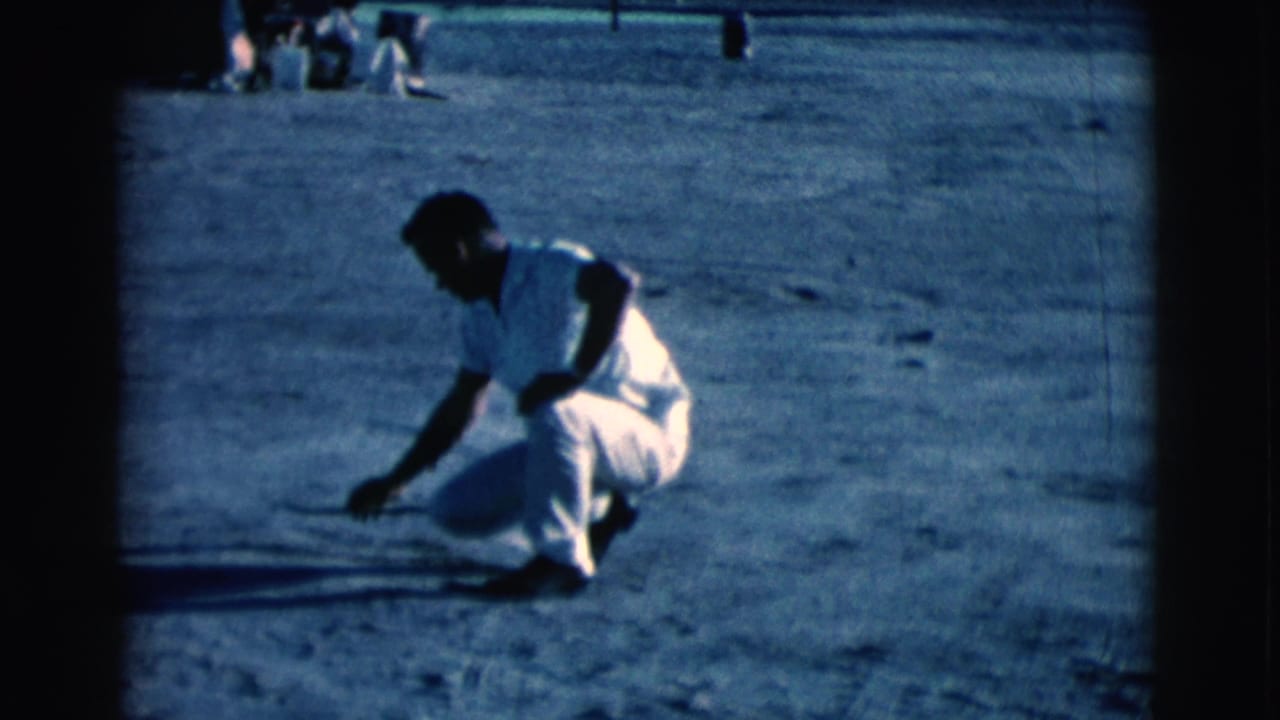 Squatting man holds a stick close to the dirt in bright daylight.