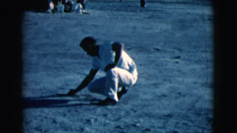 In the Clarkdale 1961 home-movie frame, the man bends over the dirt with a stick while background figures sit at the top of frame.