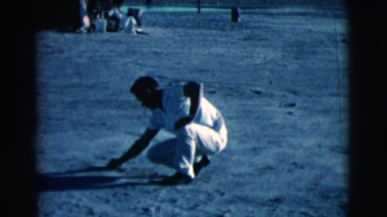 A man crouches on dusty ground and reaches toward a line or mark with a stick.