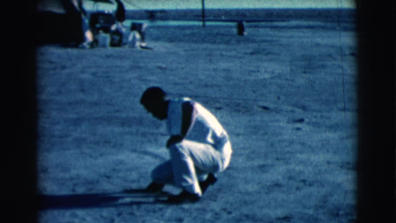 A man in light clothing crouches on open dusty ground near faint marks.