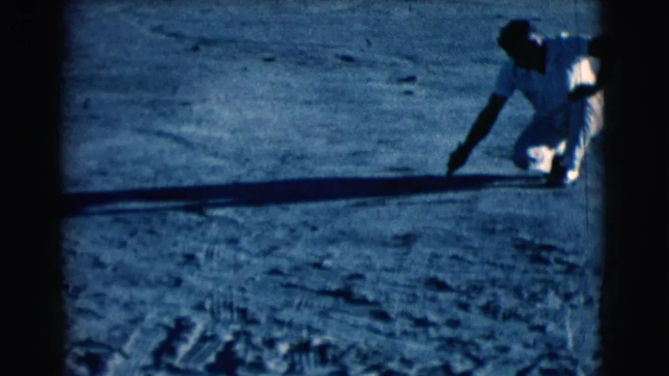 A man in light clothing crouches in Clarkdale, Arizona, extending a stick or tool toward the dry ground in a 1961 8mm frame.
