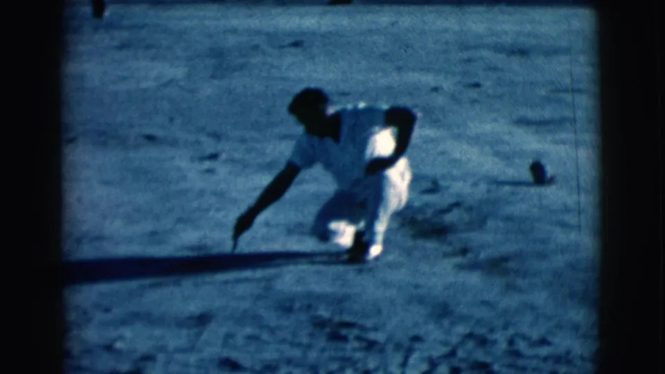 A blue-tinted 8mm frame shows the man in white crouching low and extending his arm toward the dirt in the Arizona field.