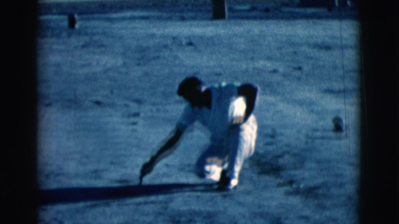 Man in light clothing crouching on sandy ground with a stick.