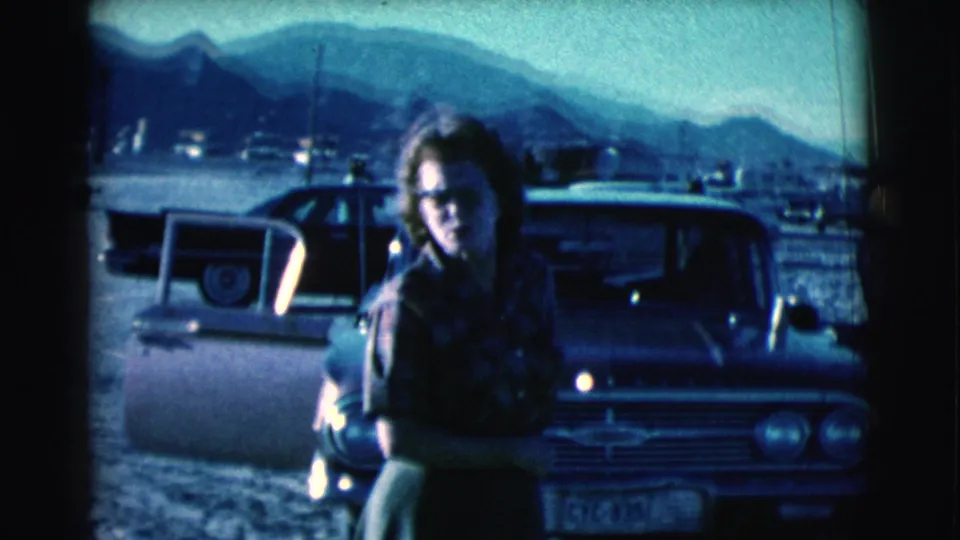 In 1961 Clarkdale, Arizona, a woman with glasses stands near classic cars with distant mountains in the background.