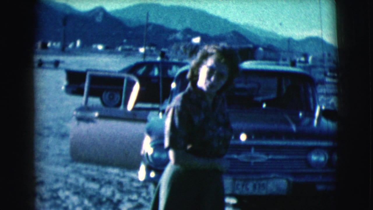 Woman standing by an open car door on sandy ground with mountains behind her.